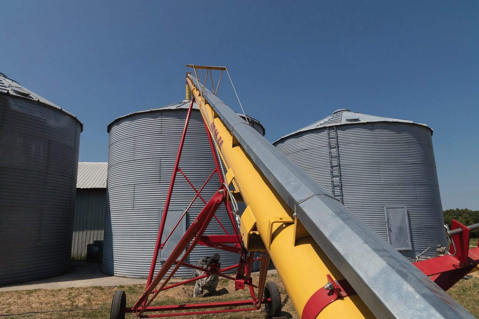 A yellow portable auger angled up toward three corrugated steel bins